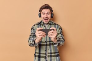 man-plays-mobile-video-game-holds-smartphone-horizontally-wears-wireless-headphones-wears-checkered-shirt-isolated-beige-people-technology-concept (1)